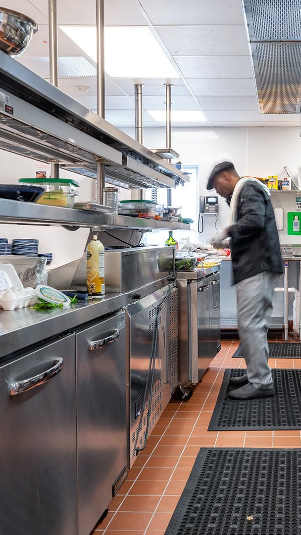 A person in a chef's uniform and cap prepares food at a stainless steel station in a commercial kitchen.