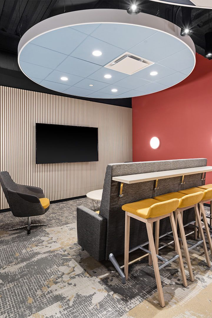 A modern office lounge with a circular light fixture, a television on a striped wall, a gray armchair, and yellow bar stools.