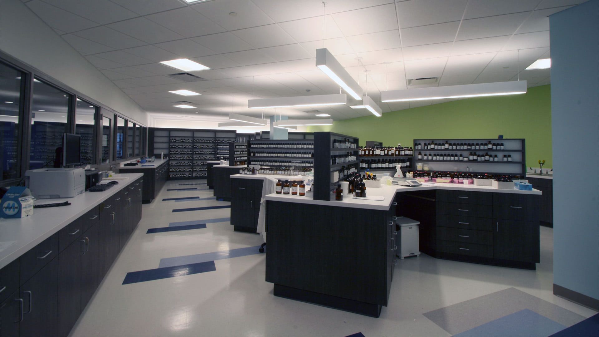 A modern pharmacy with dark cabinets, white countertops, organized shelving, and bright, hanging linear lights.