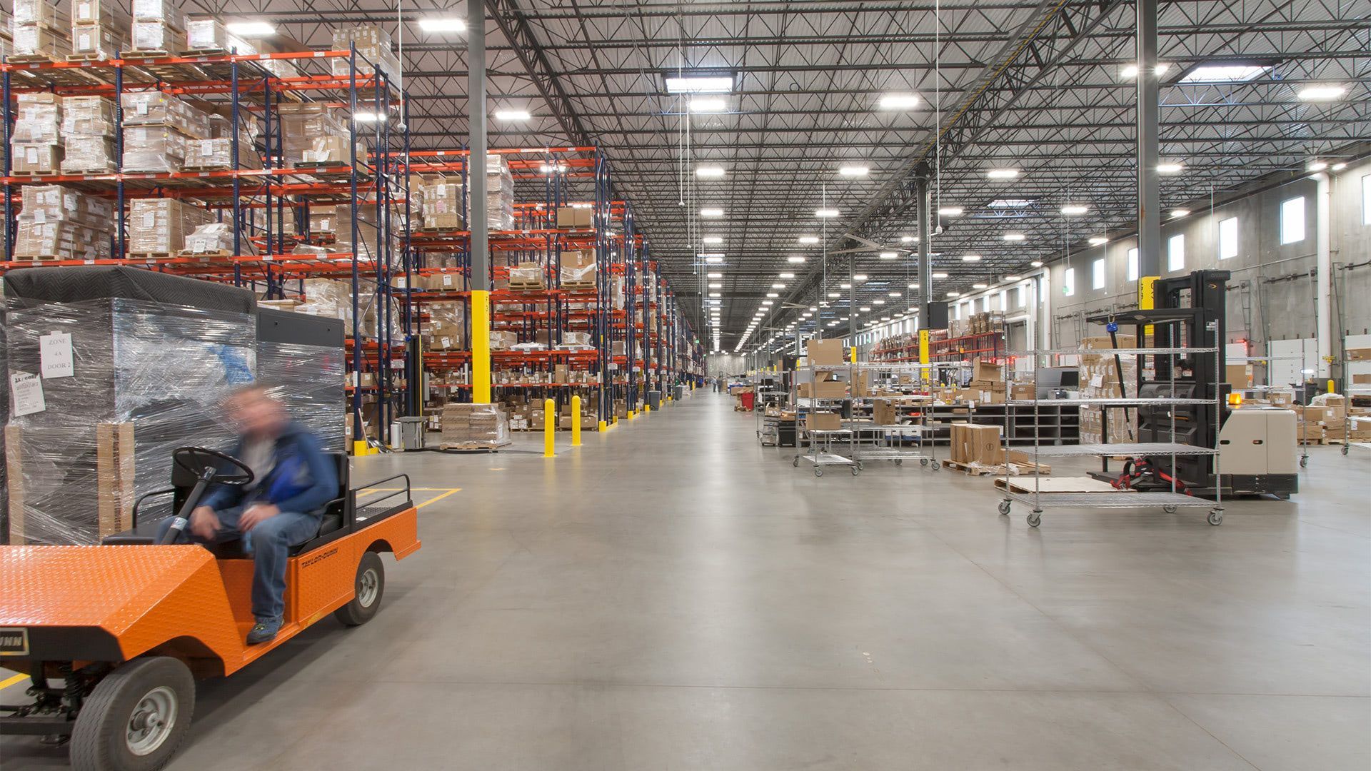 An orange utility vehicle carries a worker through a spacious warehouse filled with tall rows of storage shelving.