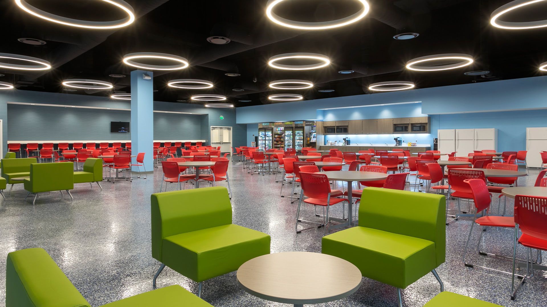 Modern cafeteria with bright green lounge chairs, red dining chairs, round tables, and circular lighting on a dark ceiling.