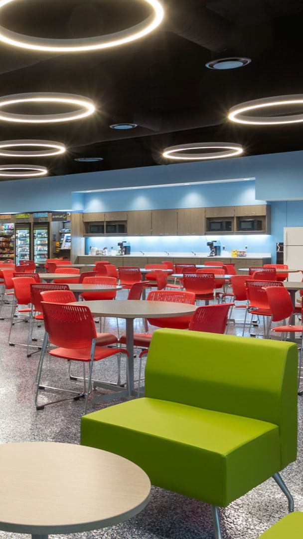A modern cafeteria with circular ceiling lights, lime green lounge chairs, and rows of red chairs at round tables.