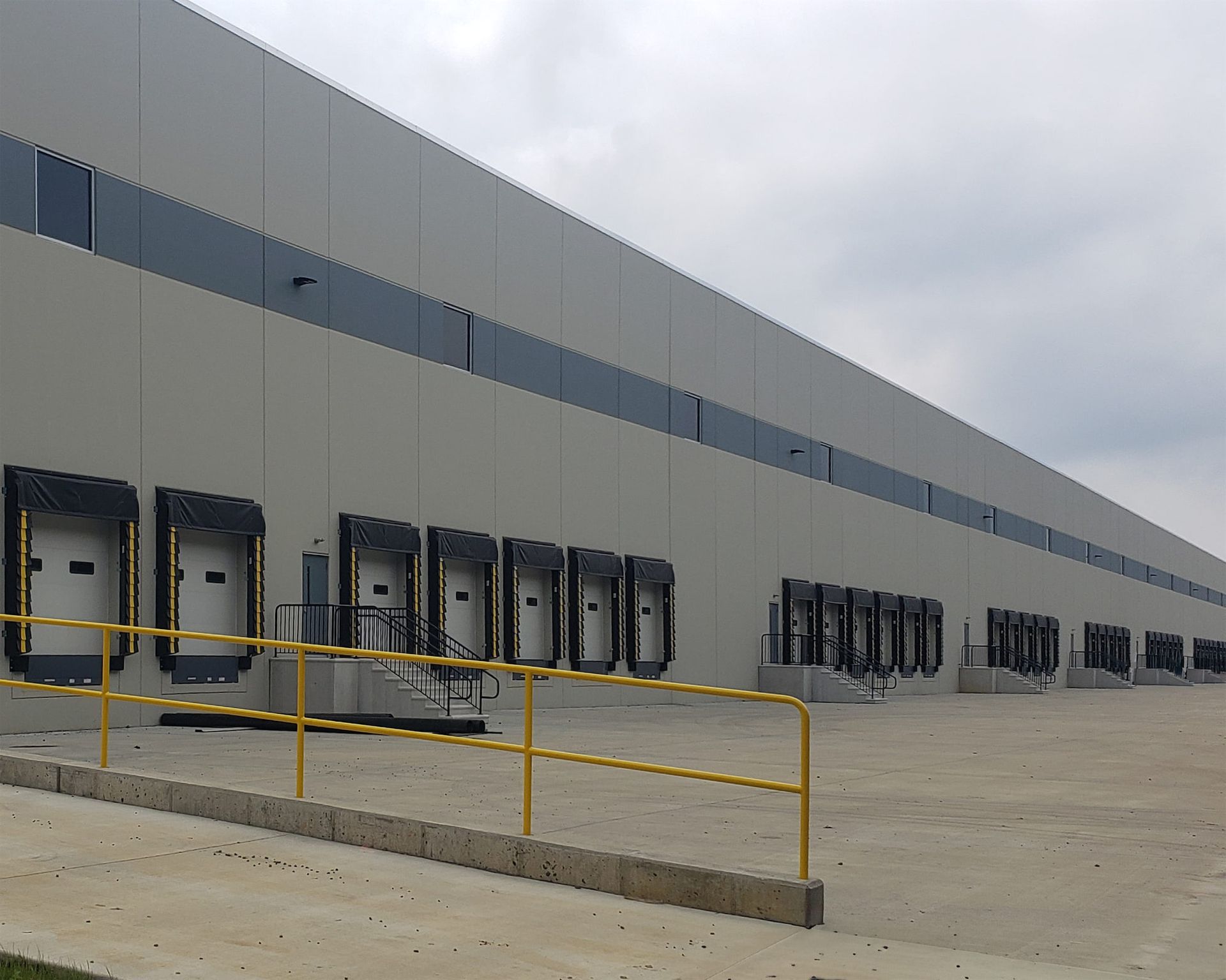 A long gray industrial warehouse exterior with multiple loading dock doors and a yellow safety railing in the foreground.