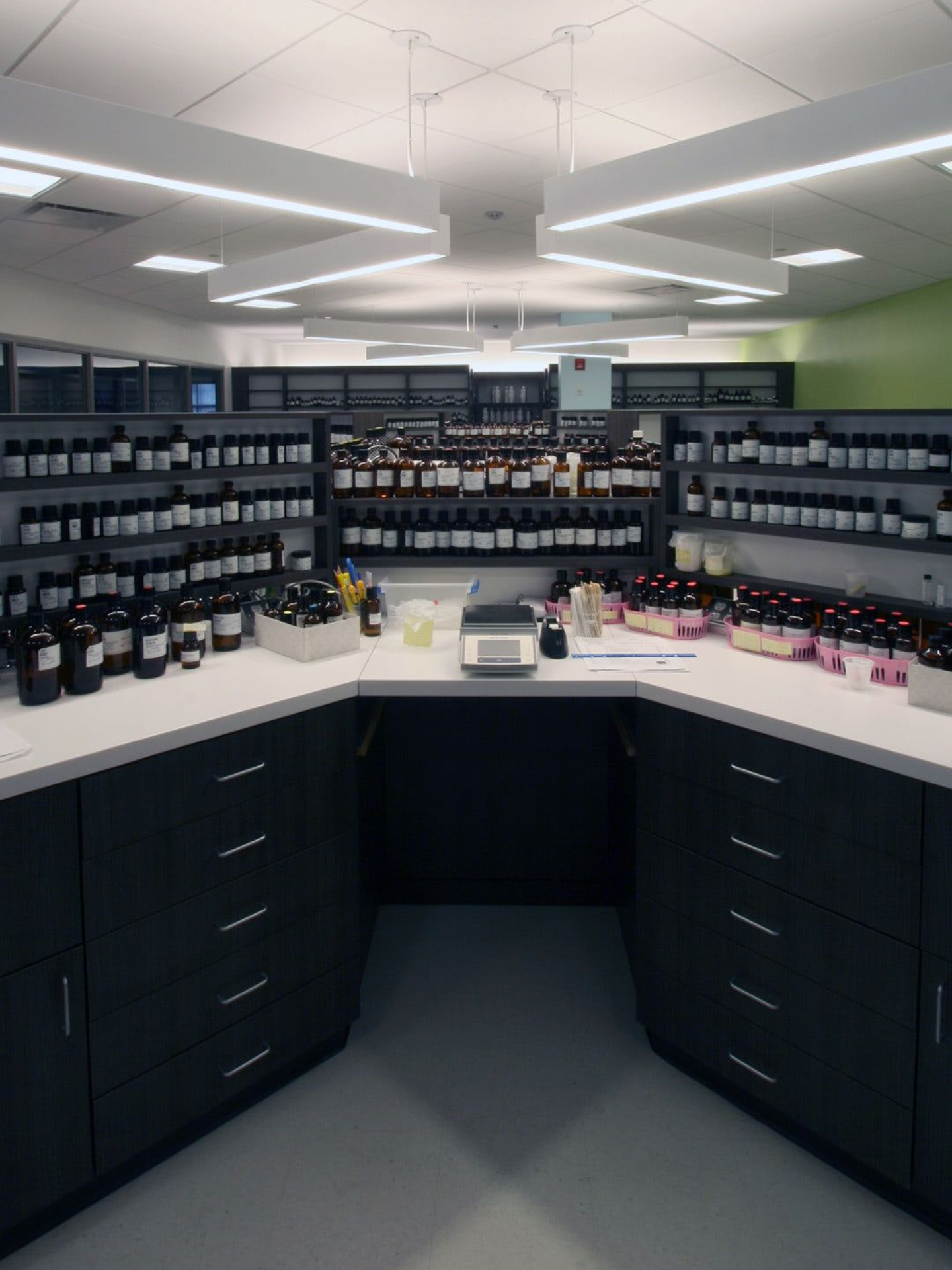 A modern laboratory workspace features a U-shaped white counter with dark cabinets and shelves filled with chemical bottles.