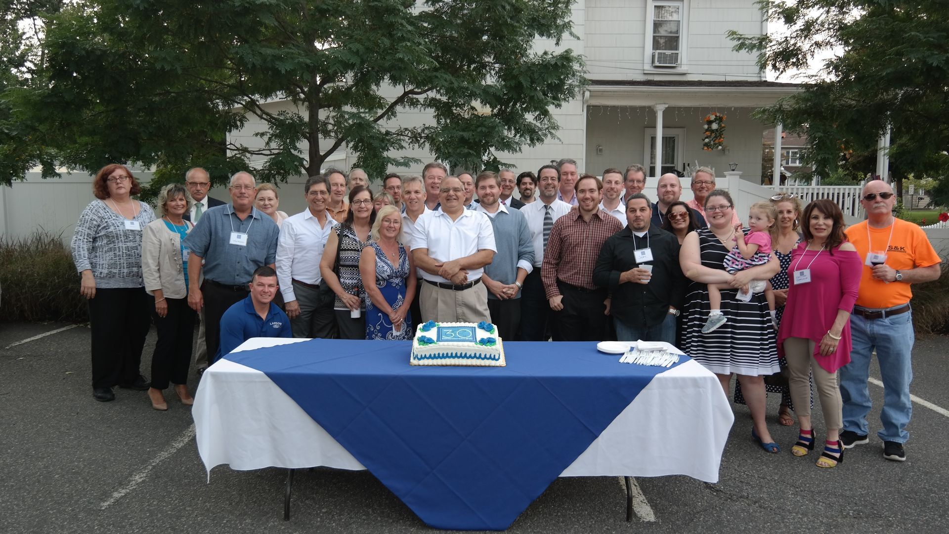 A large group poses for a photo around a table with a cake in front of a white house.