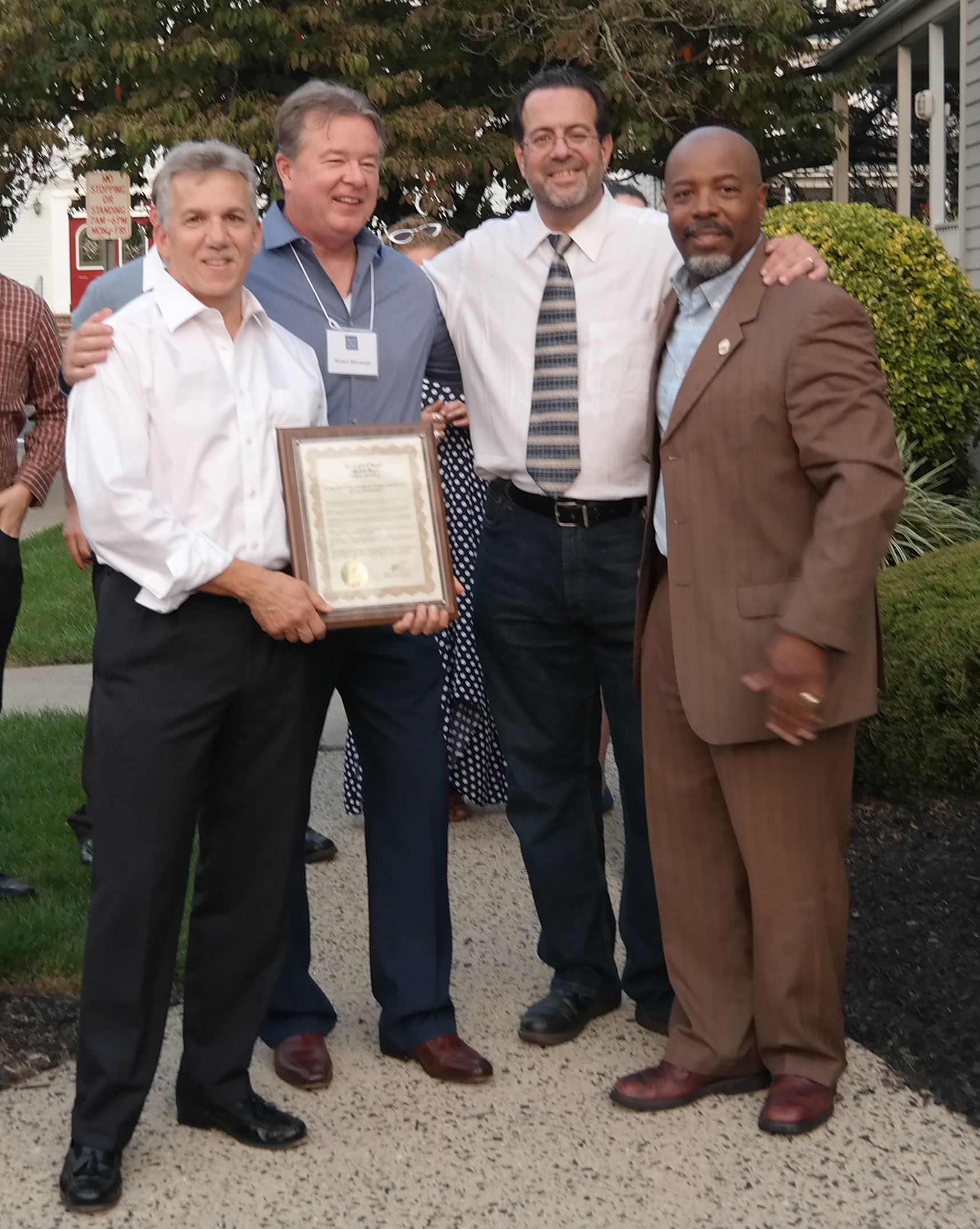 Four men standing together outdoors, smiling as two hold a framed certificate, with a park-like background.