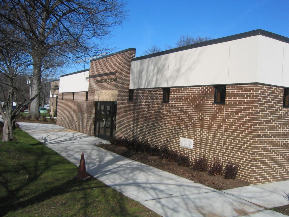 A single-story brick building with a white upper trim, a centered entryway, and a concrete walkway in a park setting.