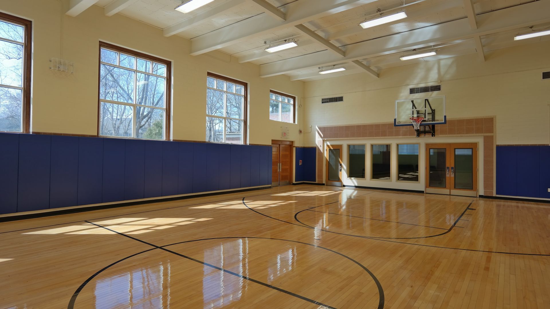 A bright, empty indoor basketball court with wooden floors, blue wall padding, large windows, and a basketball hoop.
