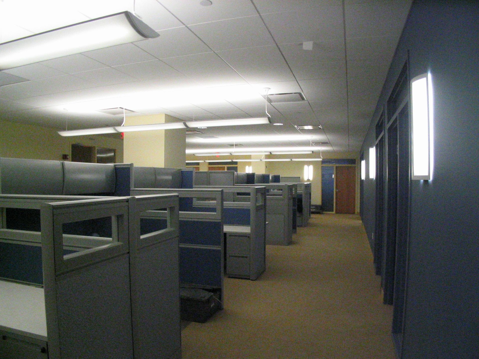 An office interior with rows of blue and grey cubicles, a carpeted walkway, and wall-mounted light fixtures.