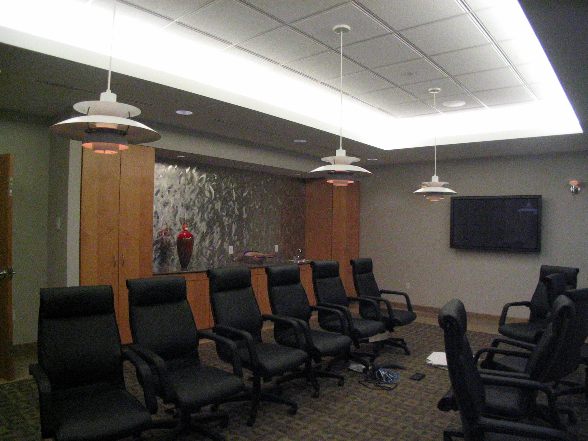 A modern conference room with a row of black ergonomic chairs, wooden cabinets, hanging light fixtures, and a TV on a wall.