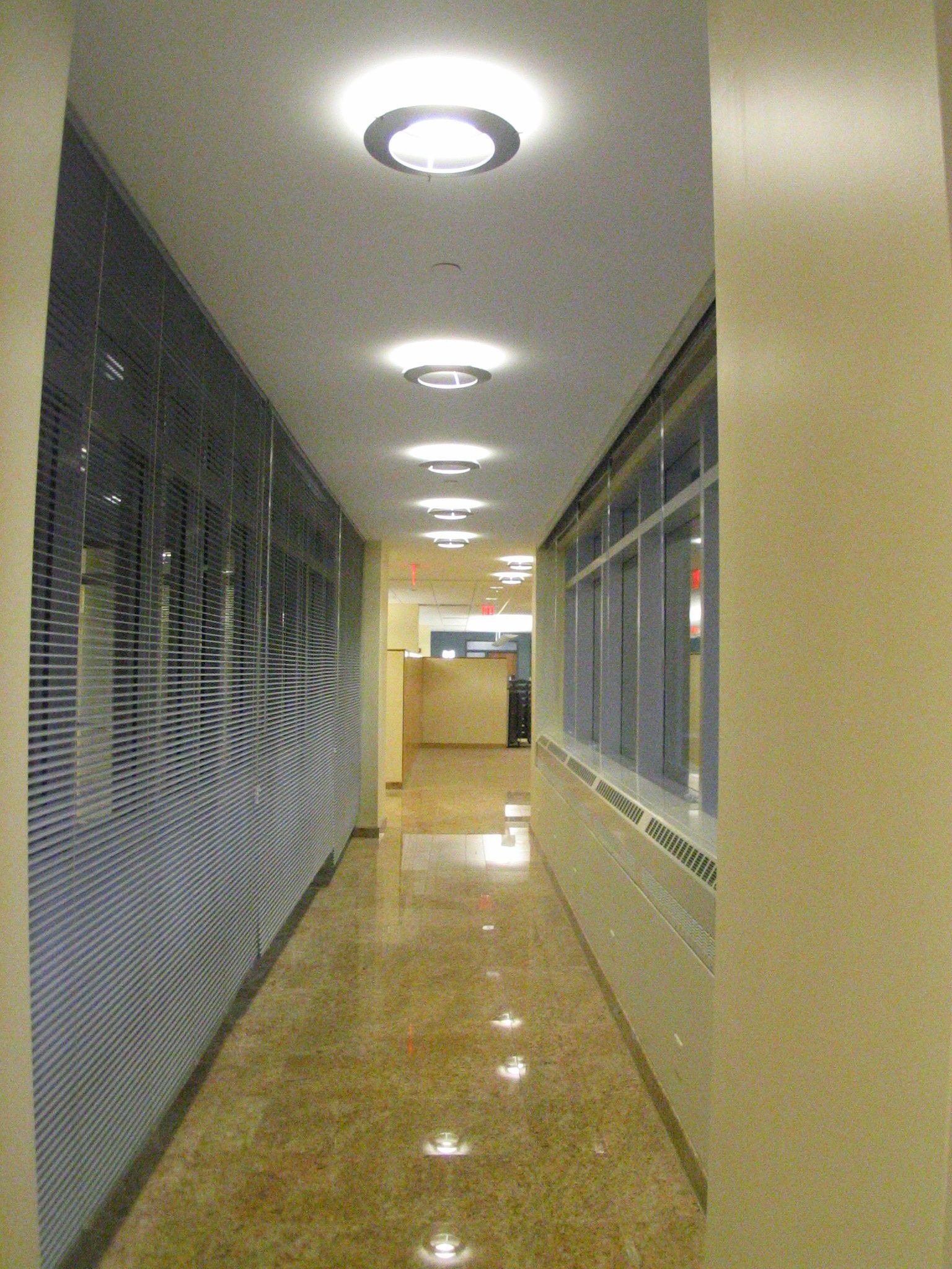 A long, indoor hallway with polished tan floors, light-colored walls, and a row of circular ceiling lights.