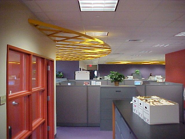 A modern office interior featuring gray cubicles, red walls, and a decorative wooden grid ceiling structure.