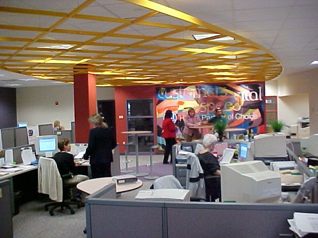 An open-plan office features a large yellow lattice ceiling structure, workstations, and a mural on the back wall.