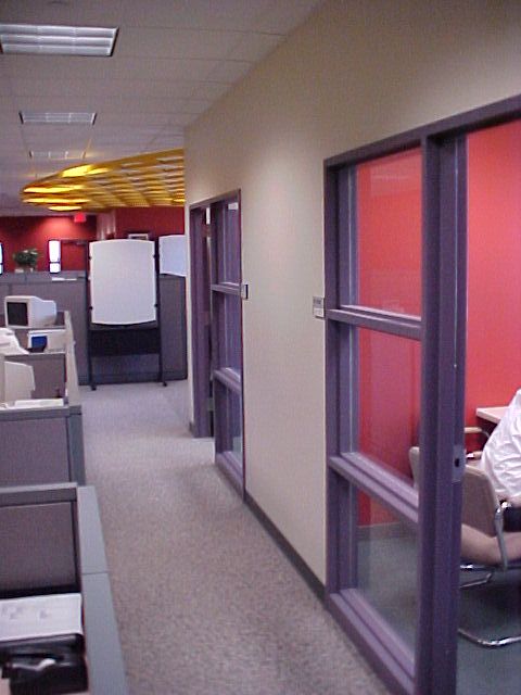 A hallway in an office featuring cubicles on the left and glass-walled, red-accented rooms on the right.