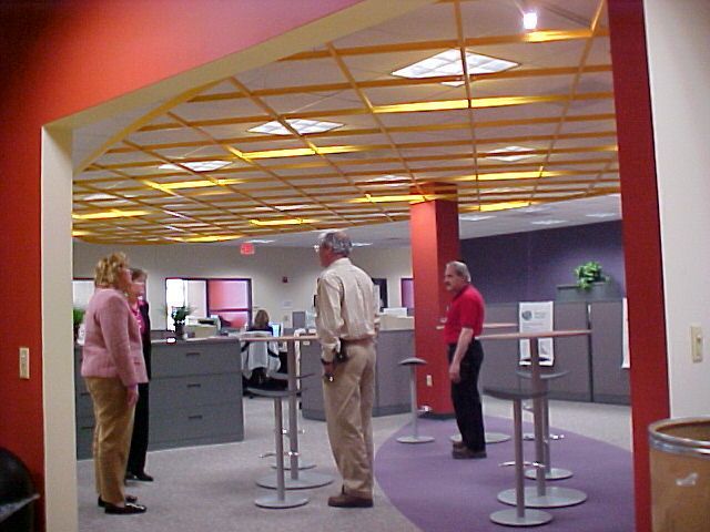 People stand and converse in a modern office with a decorative yellow grid ceiling, red accent walls, and purple flooring.