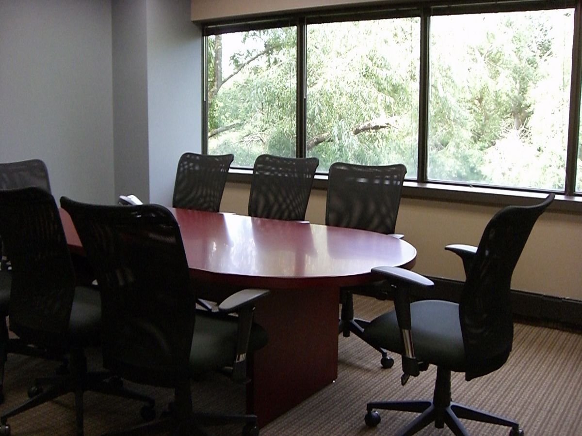 A conference room with a large, oval, reddish-brown table surrounded by six black office chairs, set before a large window.