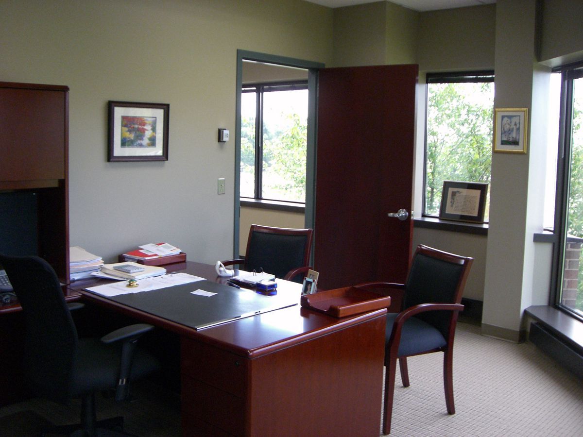 A bright office space with a large wooden desk, black chairs, framed pictures on walls, and large windows.