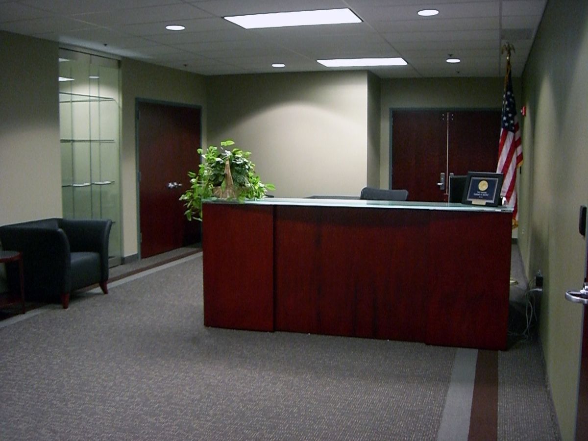 A wooden reception desk stands in an office lobby with green walls, a black armchair, and a small American flag.