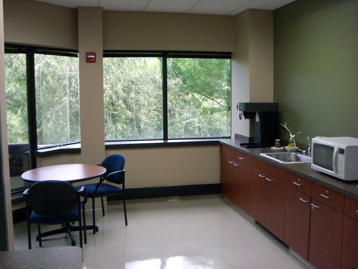 A break room with a small round table, two chairs, a counter with a sink, a coffee maker, and a microwave by a window.