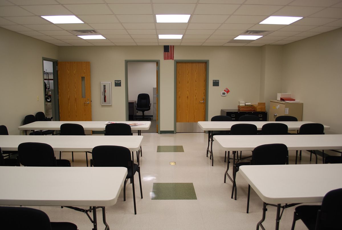 A meeting room with white tables and black chairs arranged in rows facing two wooden doors, with office lighting above.