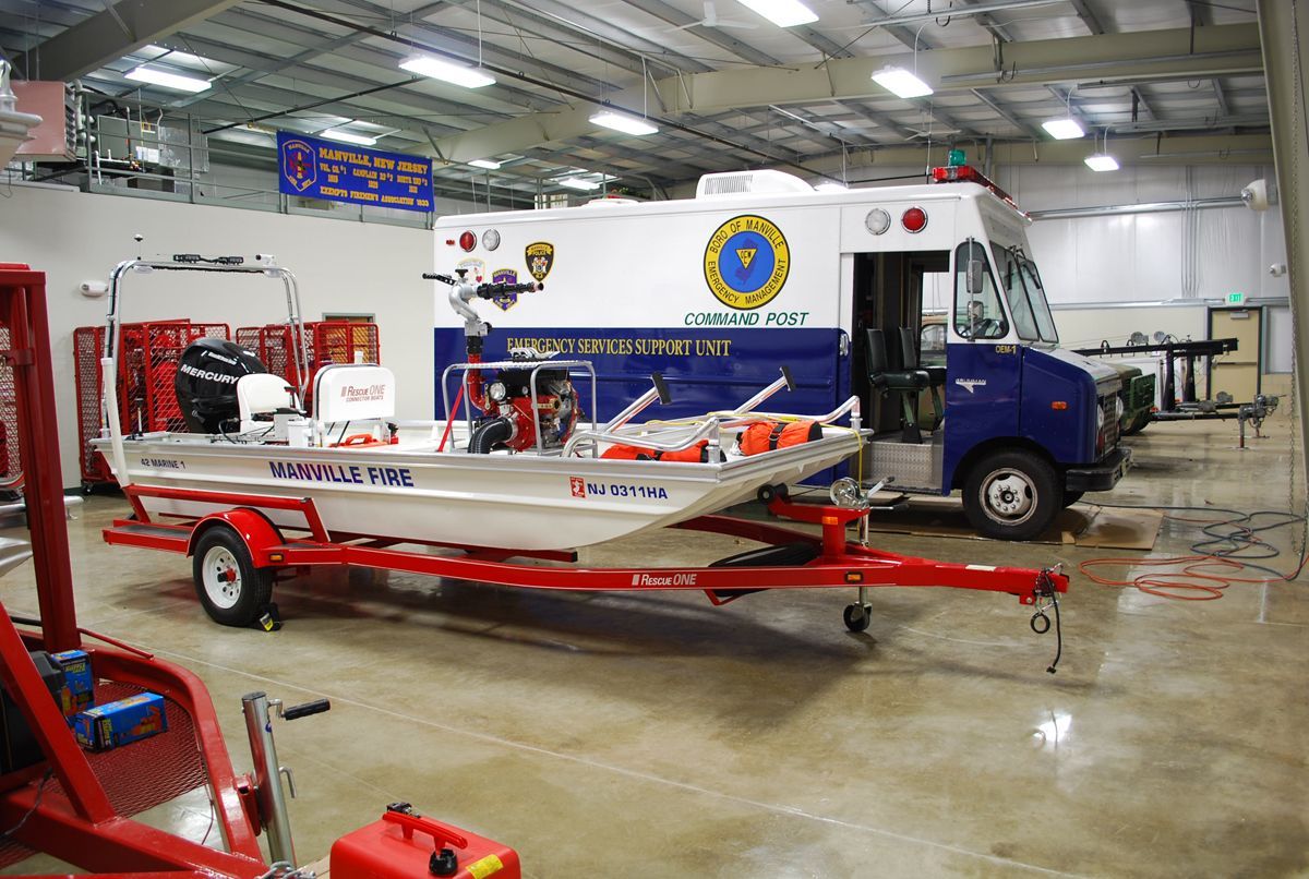 A white rescue boat on a red trailer parked inside a garage next to a blue and white emergency services truck.