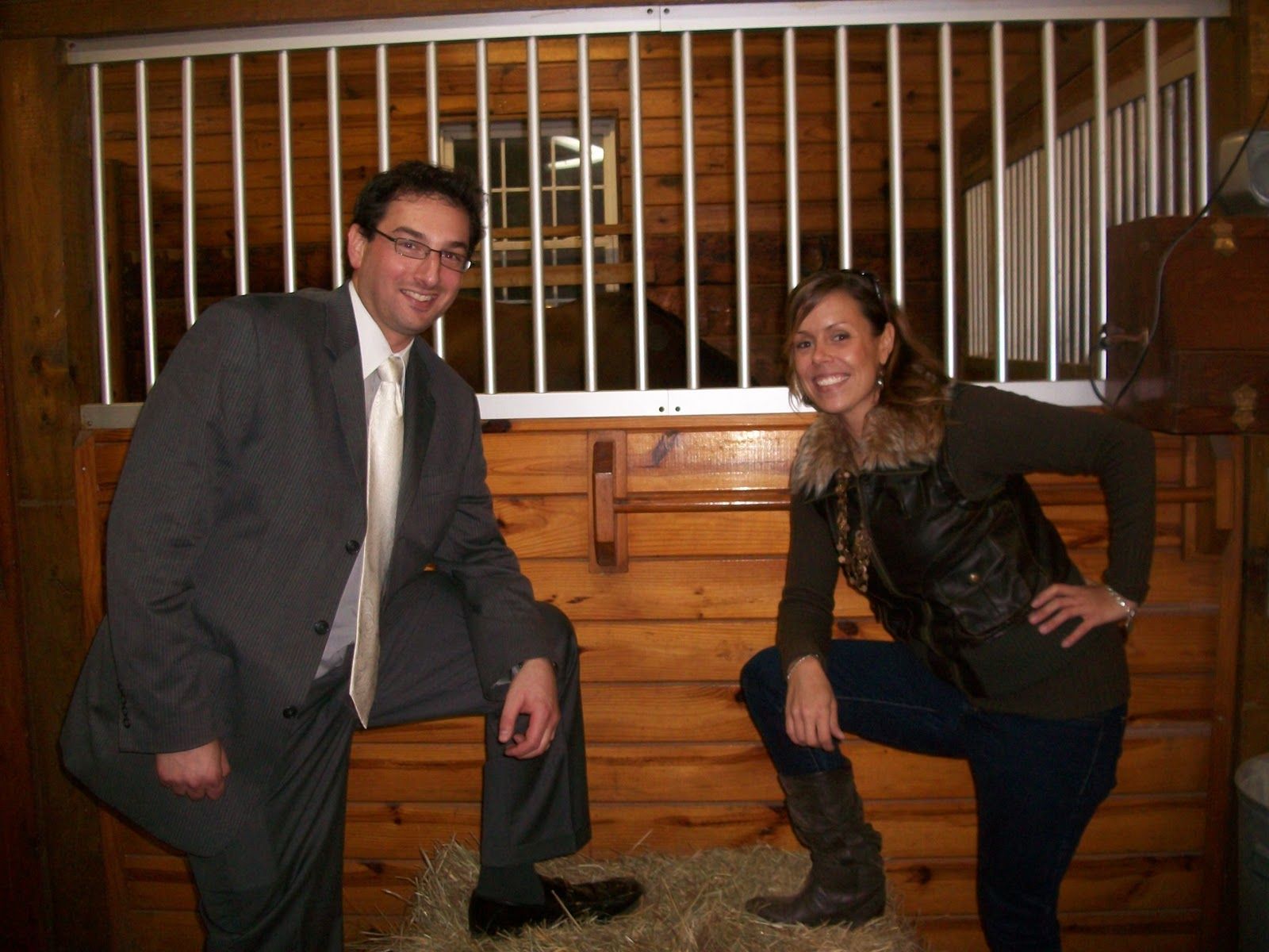 A person in a suit and a person in a vest and jeans pose in a wooden horse stable, each with a foot resting on a hay bale.