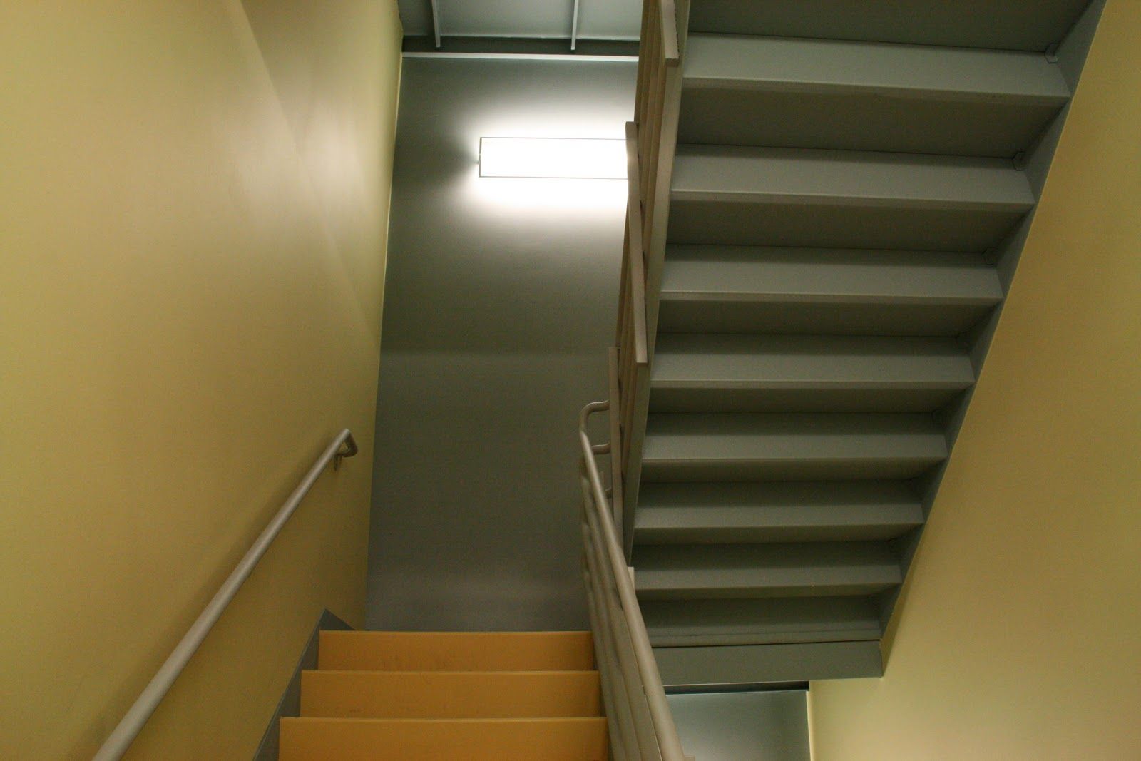 A brightly lit stairwell with yellow walls, gray concrete stairs, and a metal handrail.