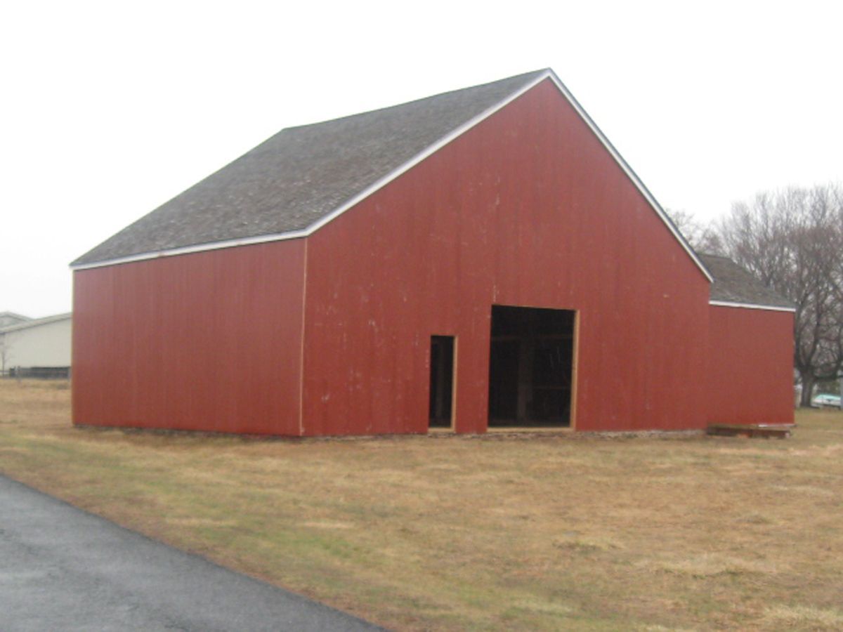 A red barn with a gray shingled roof and open doorways stands in a field of dry grass on a cloudy day.