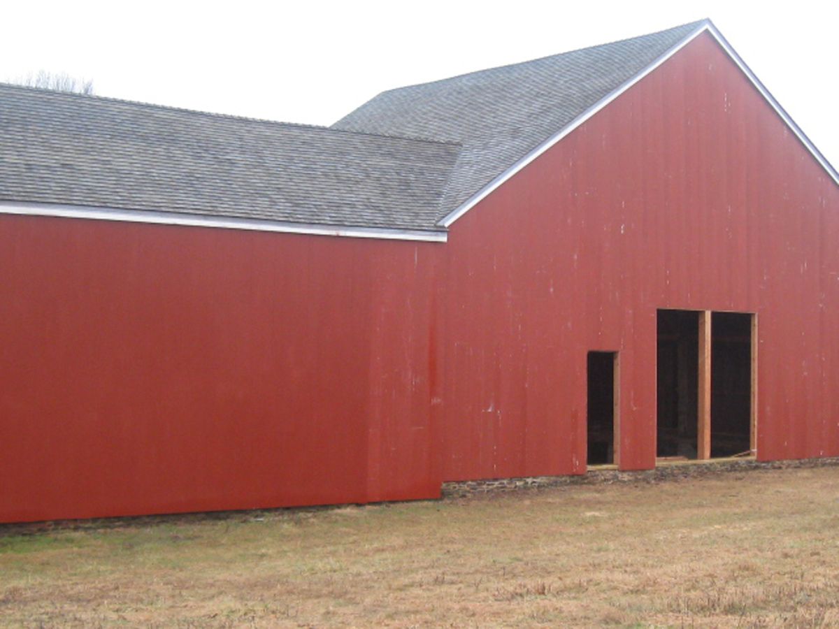 A red barn with a gray shingled roof stands on a grassy field, featuring a small doorway and a larger open entryway.