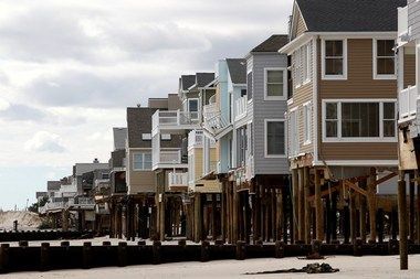 A row of multi-story beach houses elevated on wooden pilings along a sandy shore under a cloudy sky.