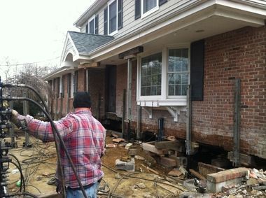 A worker operates hydraulic equipment to stabilize and lift the foundation of a brick home.
