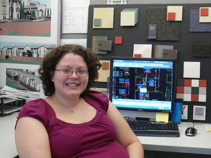 A person smiles in front of a computer monitor displaying architectural blueprints, with design samples mounted on the wall.
