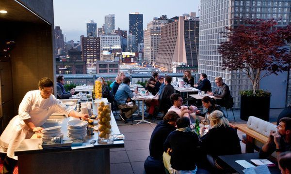 A chef prepares dishes at an outdoor rooftop restaurant overlooking a city skyline at dusk with diners at tables nearby.