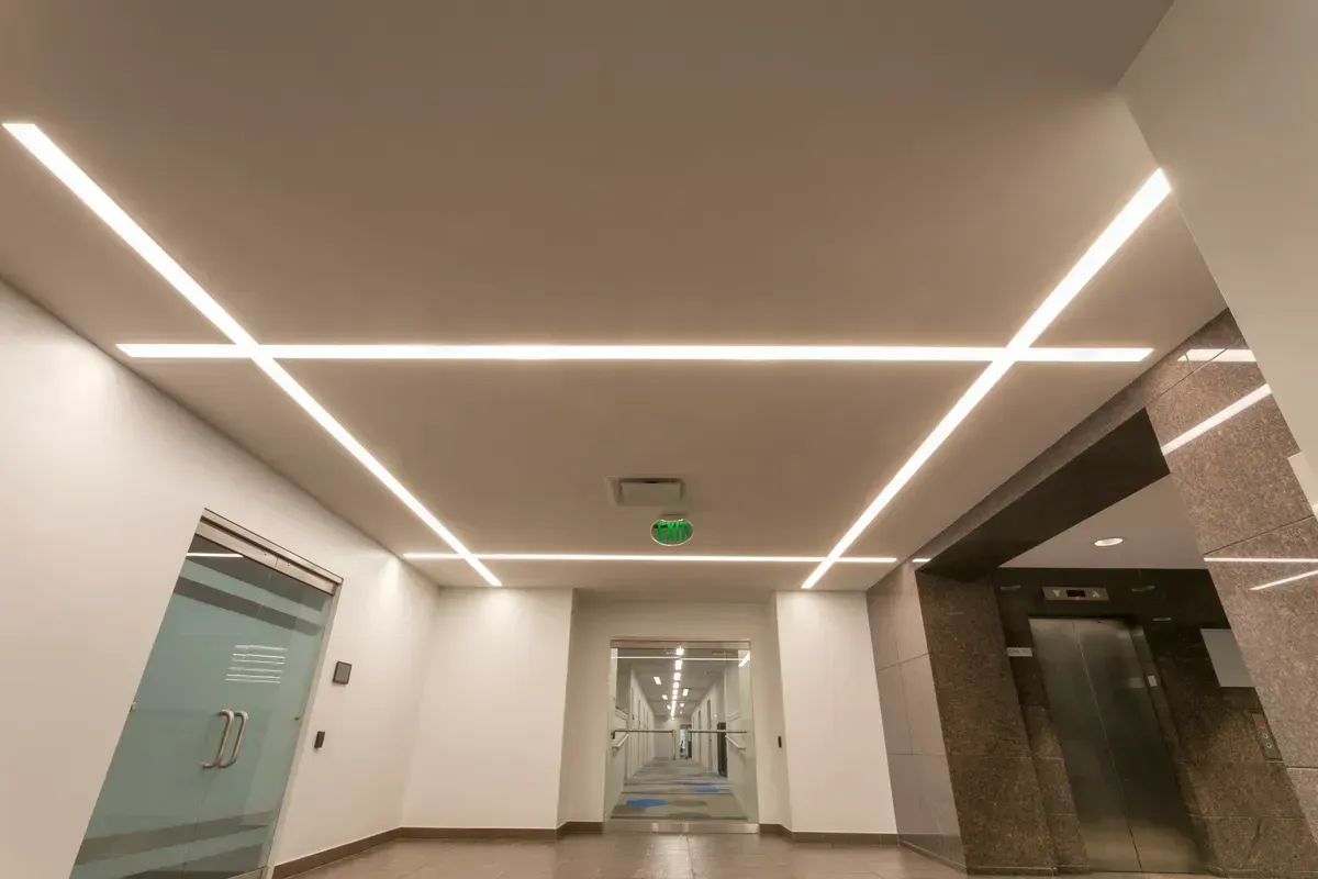 A modern office hallway with clean white walls and linear recessed lighting creating an X-pattern on the ceiling.