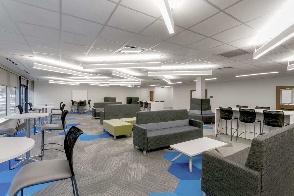 A bright, modern communal office space with modular grey sofas, tables, black chairs, and geometric blue and grey carpet.