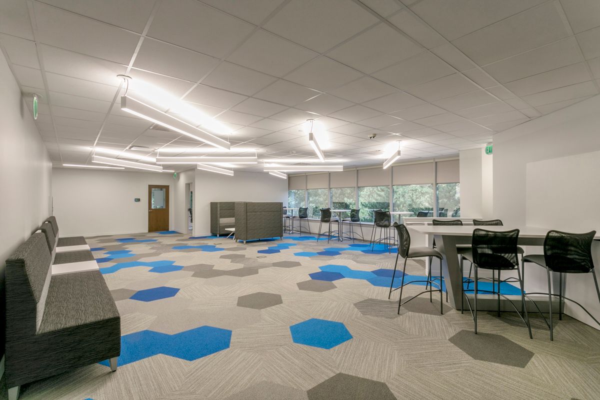 Modern office break area with gray benches, bar stools at a table, and blue and gray hexagonal floor tiles.