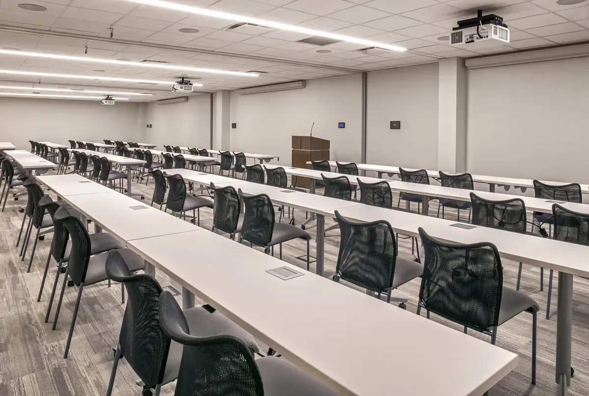 An empty meeting room with long rows of tables and black mesh chairs facing a podium.