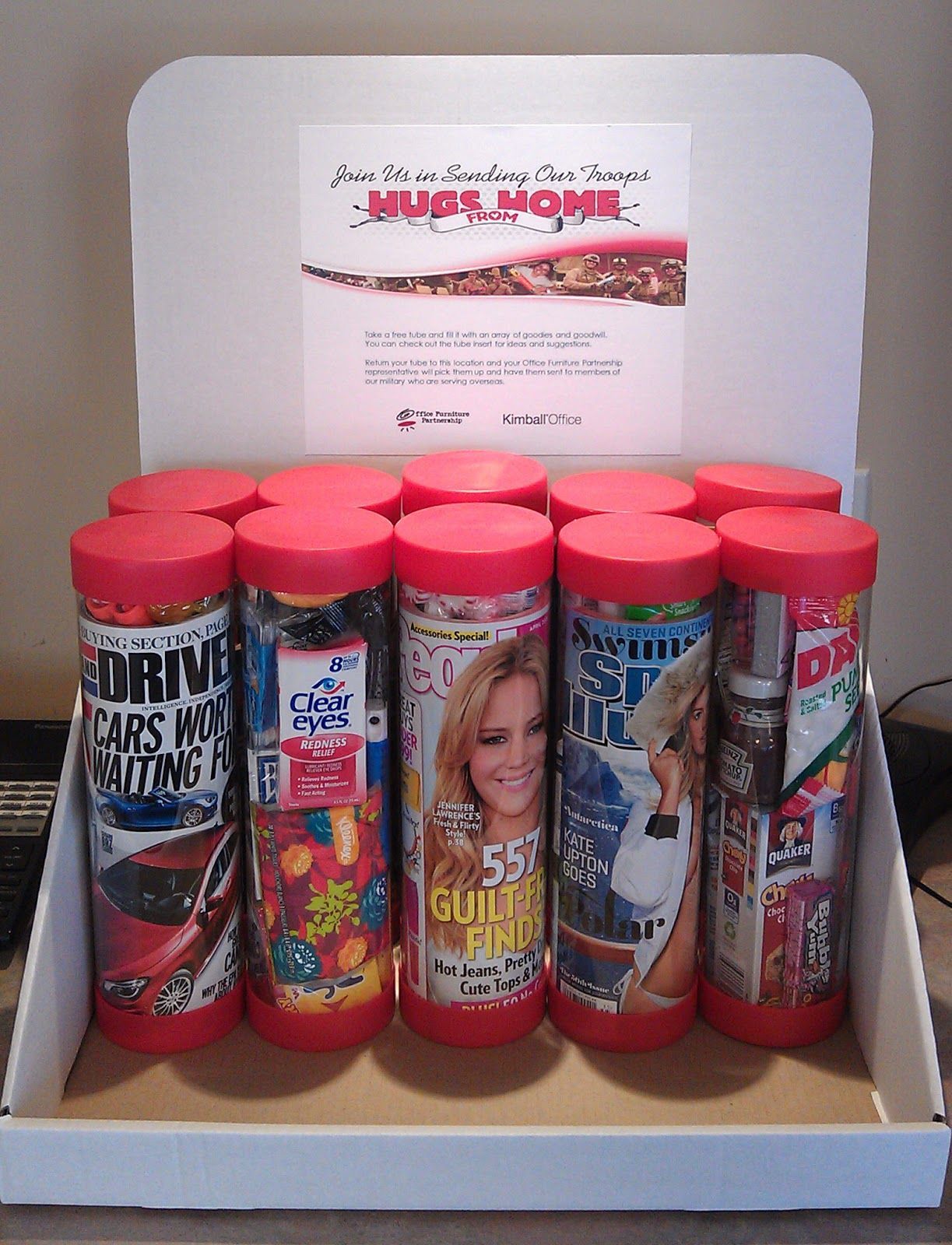 A white display box holds ten clear tubes with red lids, each filled with rolled magazines or paper inserts.