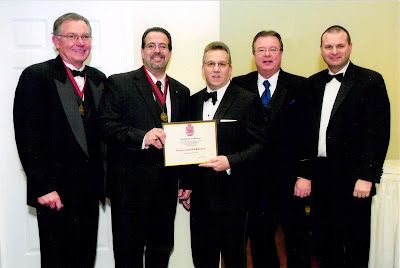 Five men in black tuxedos stand in a row; the center two hold a framed certificate at an indoor formal event.