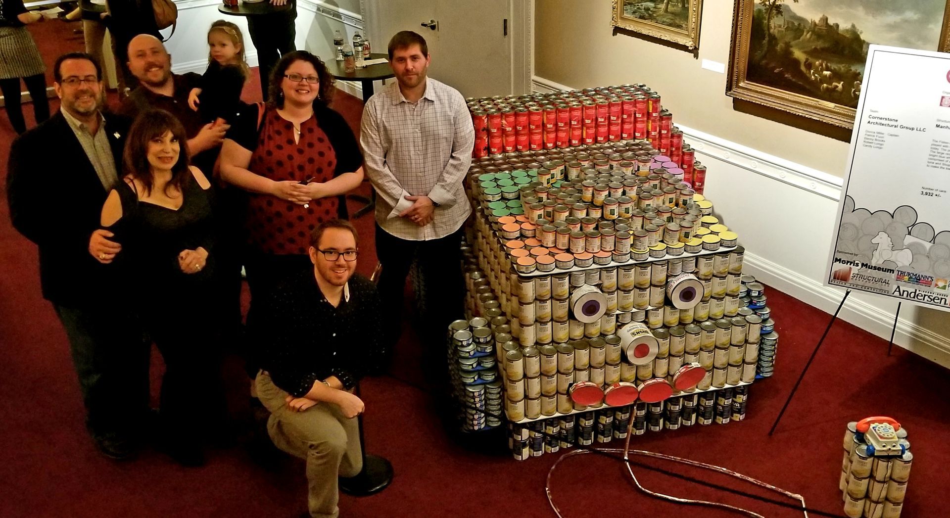 A group poses beside a large, multi-colored structure built from stacked food cans in an indoor setting.