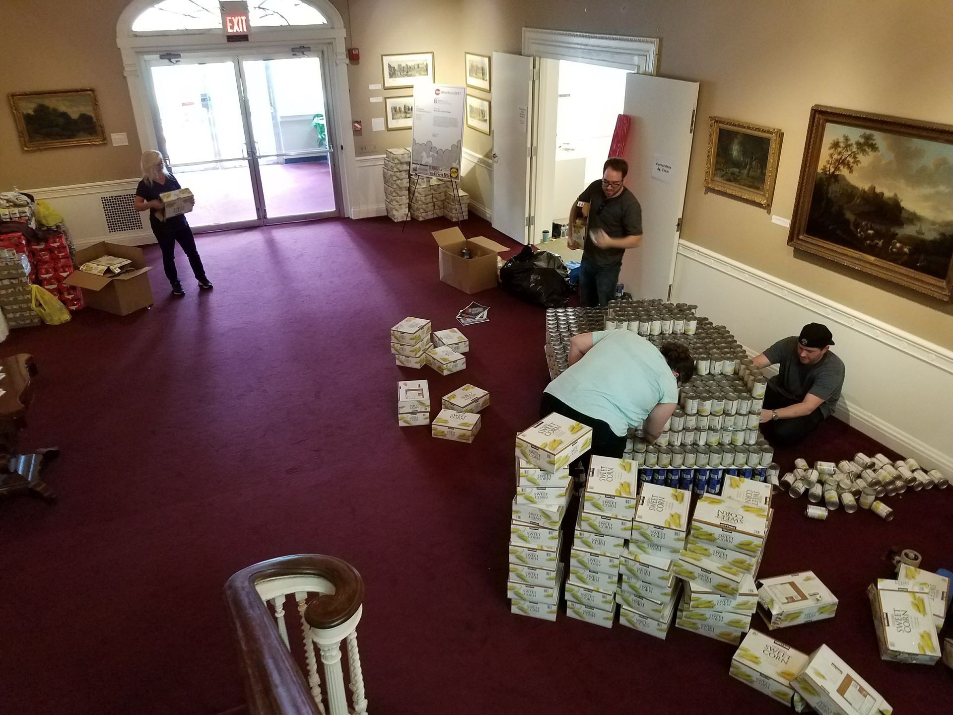 People in a carpeted room organize and stack cardboard boxes of canned goods, possibly for a food drive or donation effort.