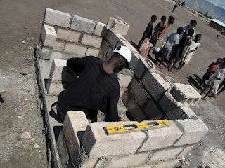 A worker in a hard hat builds a cinder block structure while a group of people stands nearby in an outdoor area.