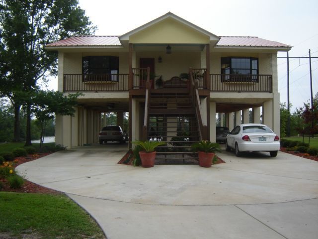 A yellow stilt house with a metal roof, front stairs, and a concrete driveway with two cars parked underneath.
