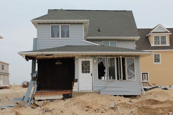 A damaged, light-blue house sitting on sand, with debris, missing wall siding, and broken windows following a storm.