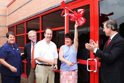 Five people gathered for a ribbon-cutting ceremony outside a brick building with large red-framed glass doors.