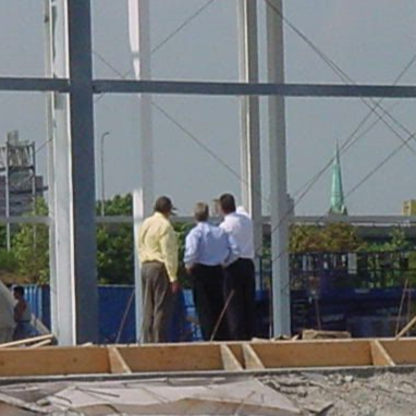 Three people in business casual attire stand at a construction site, observing the steel framework of a building.