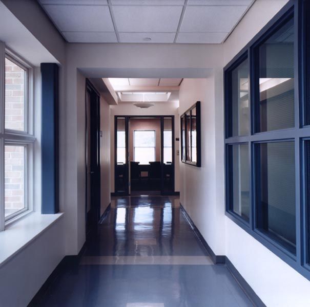 An empty office hallway with reflective floors, side windows, and a doorway leading to a room with a window at the end.