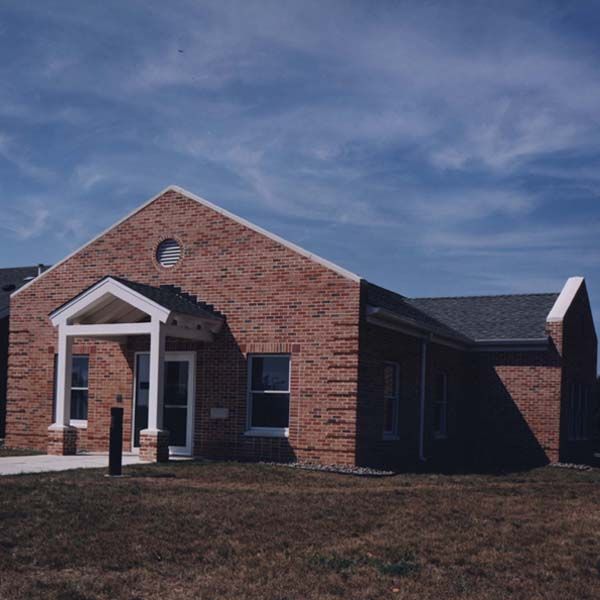 A single-story brick building with a white gabled entryway under a blue sky, set against a grassy field.