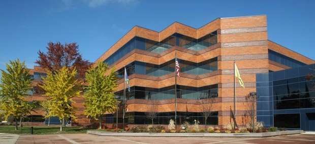 A four-story, zigzag-shaped brick office building with dark windows, surrounded by trees with autumn foliage.