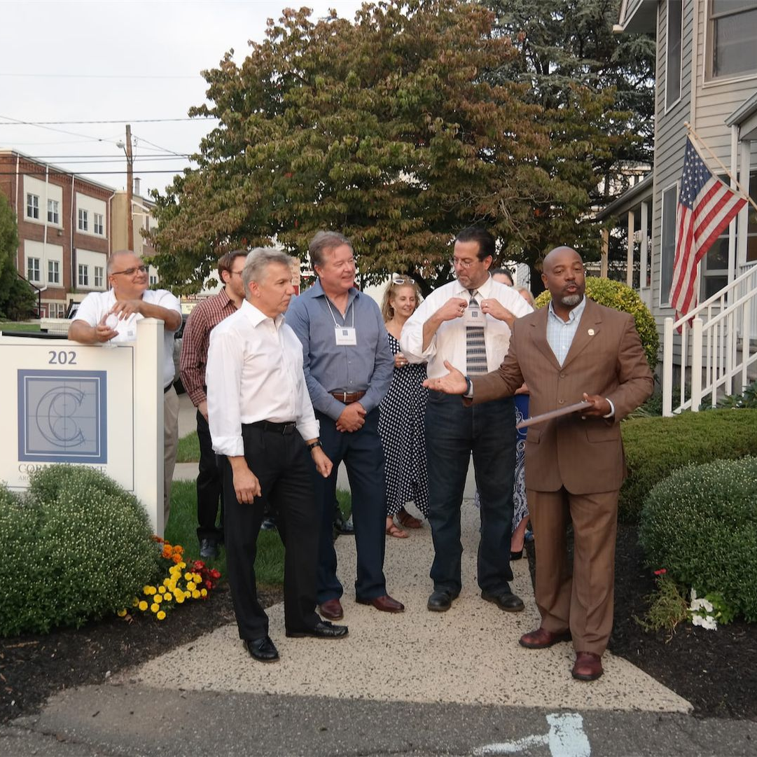A group of people standing outside a building with an American flag, with one person speaking while holding a plaque.